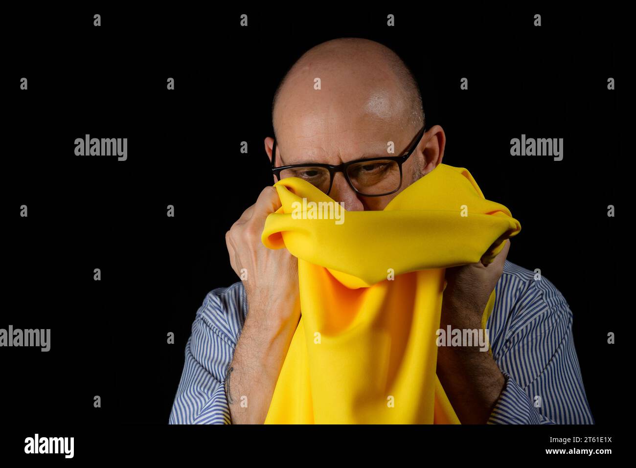 Homme chauve, barbu avec des lunettes tenant un tissu de couleur jaune au-dessus de sa tête sur fond noir. Banque D'Images