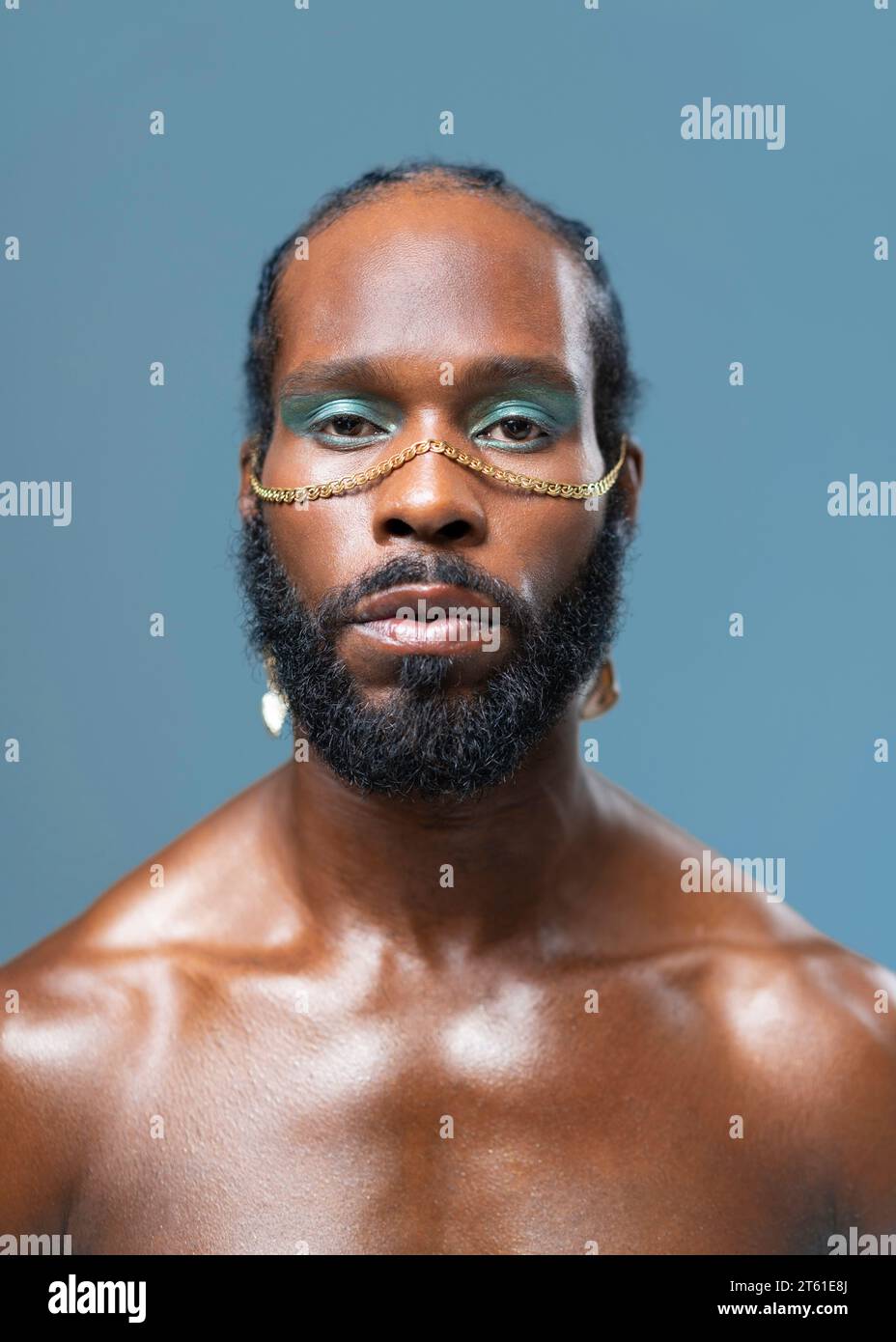 Black gay se tient isolé sur fond bleu et regarde dans la caméra avec l'expression sérieuse sur le visage. Portrait rapproché de beaux adultes transgenres avec des fards à paupières bleus et un accessoire doré sur le visage Banque D'Images
