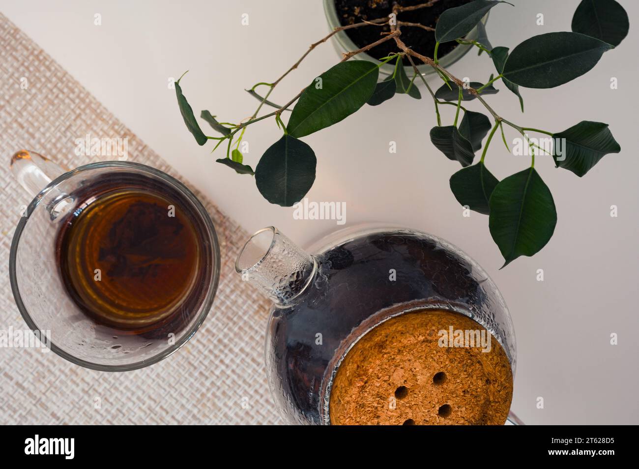 Thé noir dans une tasse en verre et théière en verre sur une serviette et plante verte sur une table blanche du haut Banque D'Images