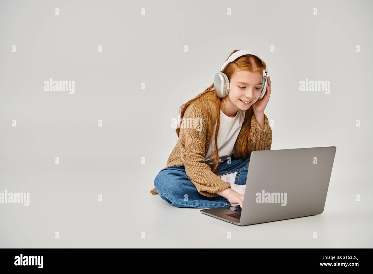 jolie petite fille joyeuse en tenue tendance d'hiver sur le sol avec casque regardant ordinateur portable, mode Banque D'Images
