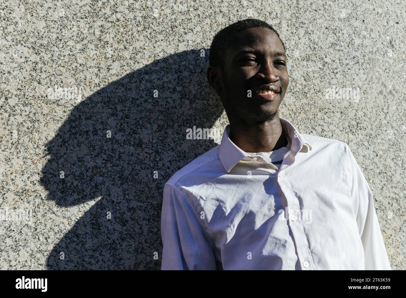Heureux jeune homme appuyé contre un mur texturé, projetant une ombre distincte avec un sourire contenu Banque D'Images