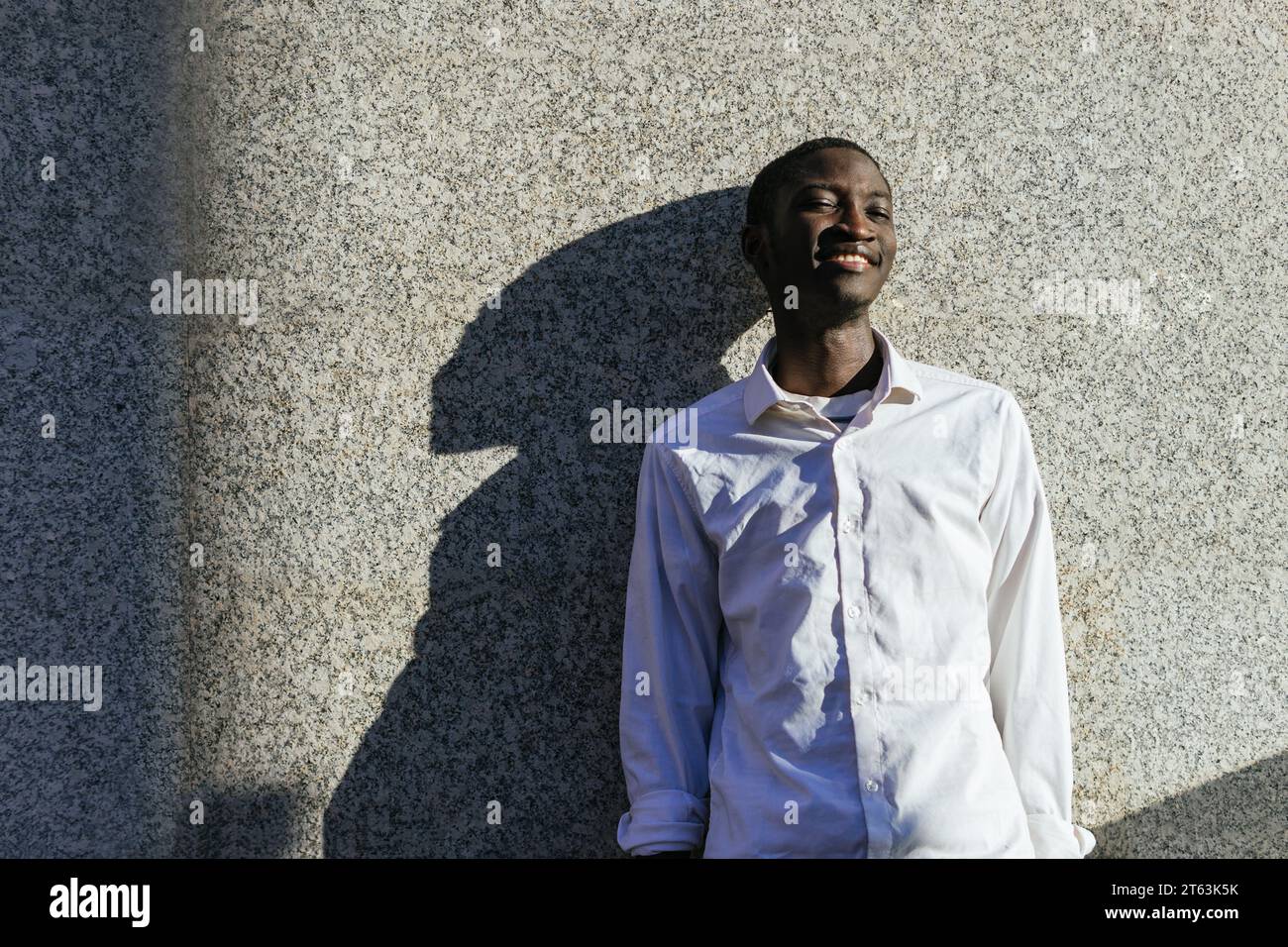 Heureux jeune homme appuyé contre un mur texturé, projetant une ombre distincte avec un sourire contenu Banque D'Images