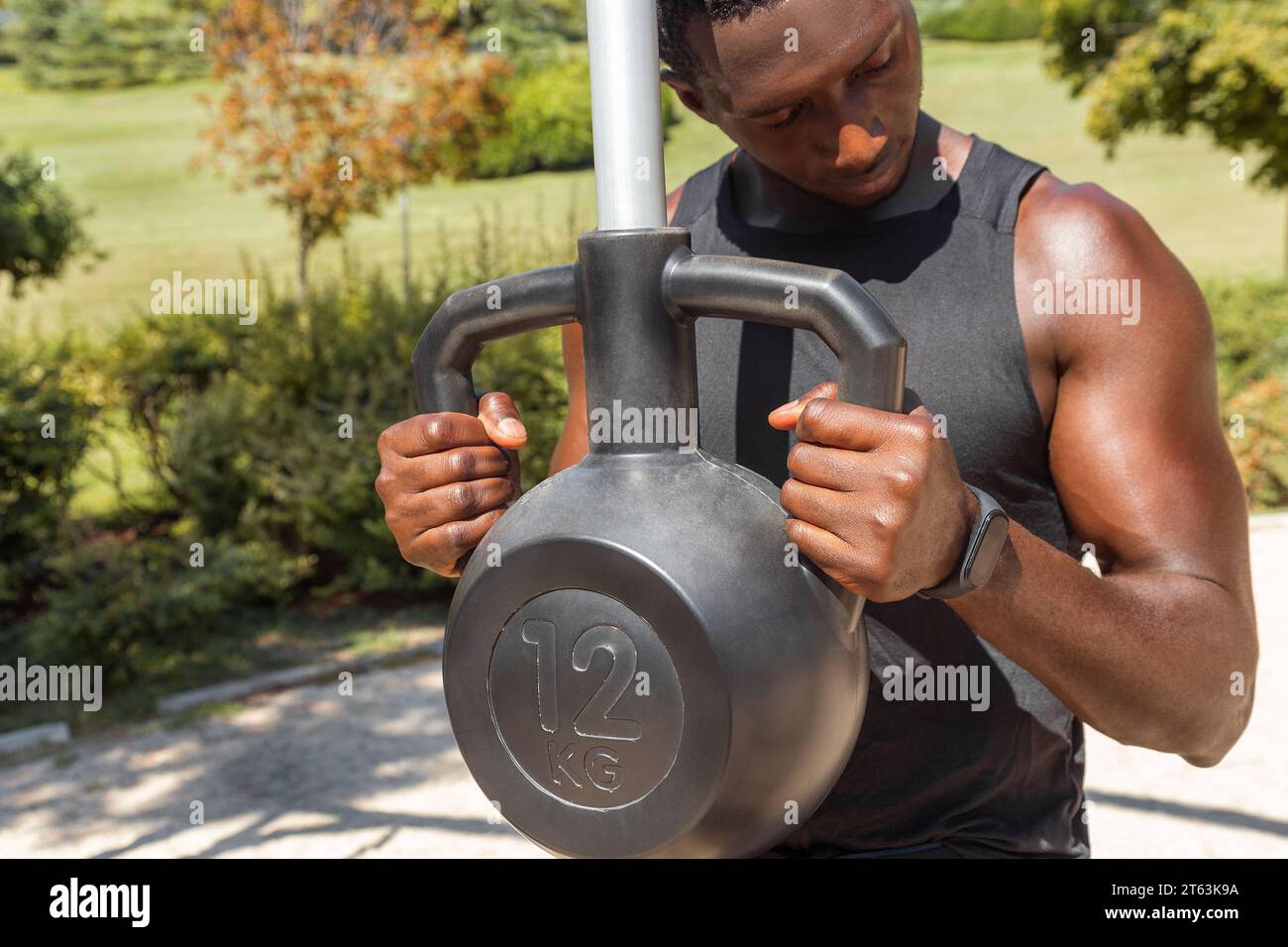 Homme noir concentré en sportswear soulevant une kettlebell de 12 kg à l'extérieur avec de la verdure en arrière-plan Banque D'Images