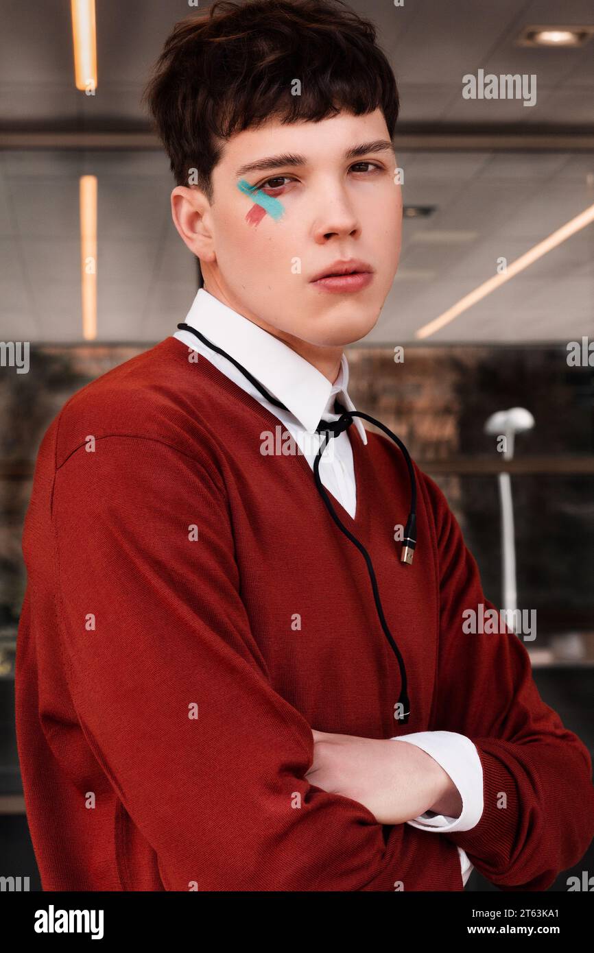 Jeune modèle masculin gen-z avec peinture pour les yeux colorée et maquillage du visage en pull rouge avec un câble de connexion attaché à son cou dans un cadre moderne. Banque D'Images