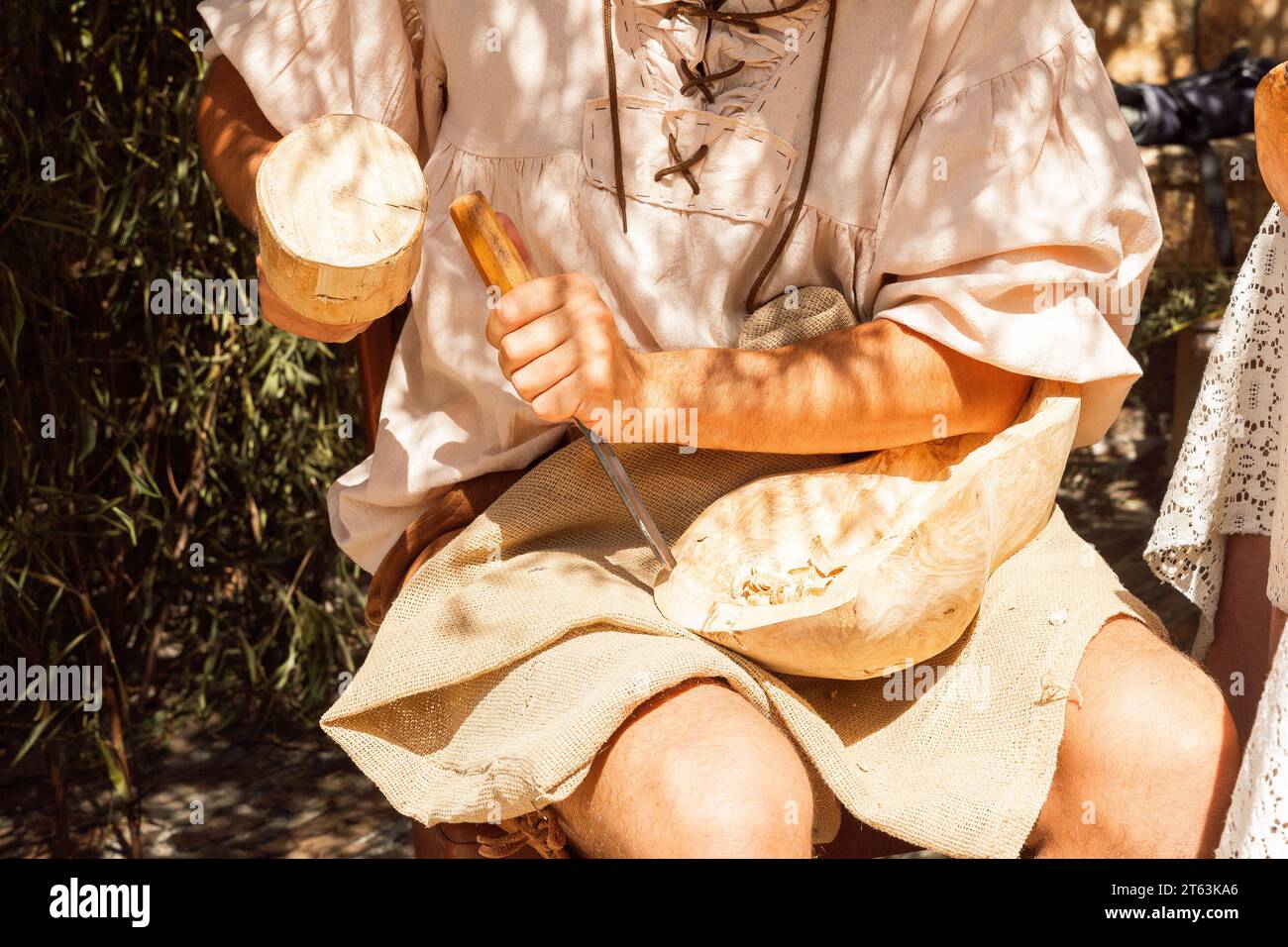 Un artisan médiéval anonyme en tenue traditionnelle sculpte habilement un bol en bois à l'aide d'outils à main assis au milieu de la verdure Banque D'Images