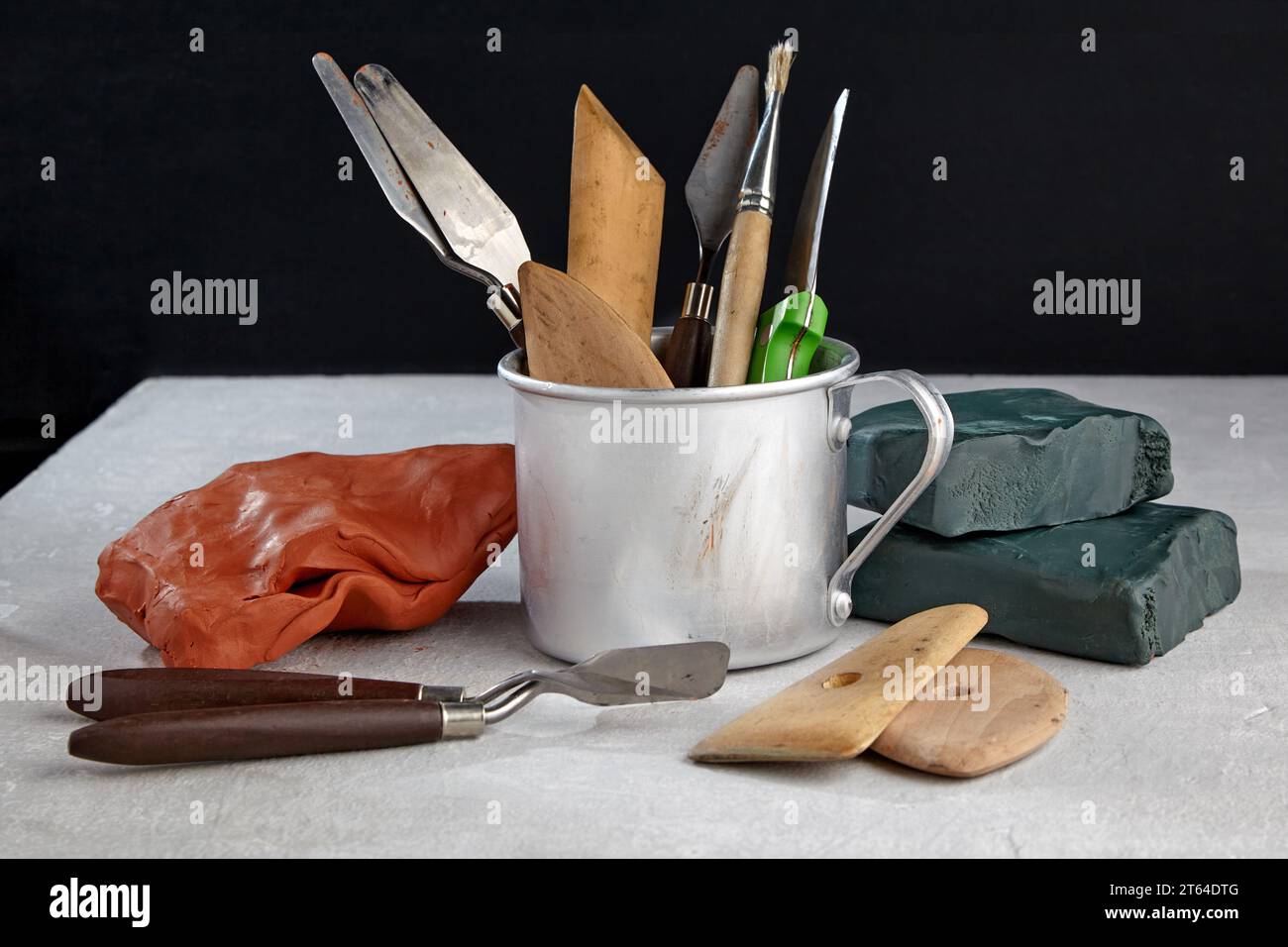 Outils de modélisation à partir de plasticine et argile dans une tasse en métal et des morceaux d'argile et de plasticine sur une table en béton blanc sur fond noir. Banque D'Images