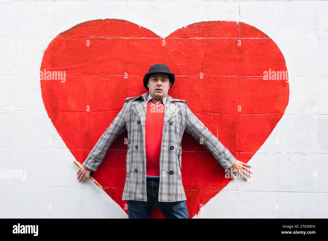 Homme émotionnel dans un chapeau et manteau debout contre un mur blanc sur un fond de coeur rouge. Concept d'amour et de relation Banque D'Images