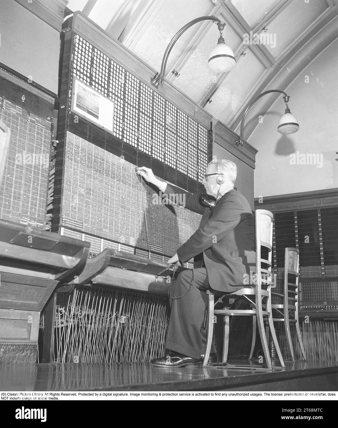 Central téléphonique. Un homme est assis à un standard manuel. Tous les appels entrants et sortants doivent être connectés et il le fait en branchant le cordon et la fiche à la bonne connexion. Le standard téléphonique est un objet de musée du tournant du siècle 1800-1900 et l'homme démontre sa fonction. Suède 1953. Kristoffersson réf. BM23-6 Banque D'Images