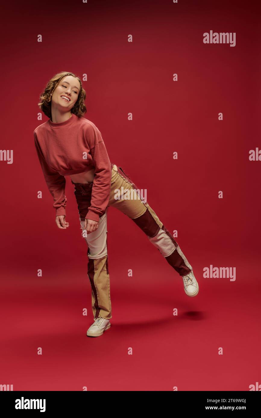 style personnel, jeune fille positive en pantalon patchwork et manche longue courte posant, fond rouge Banque D'Images