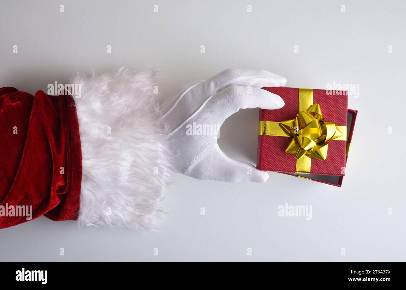 Main du Père Noël avec gant blanc découvrant la boîte cadeau rouge avec noeud doré habillé de son costume rouge traditionnel sur la table blanche. Vue de dessus. Banque D'Images