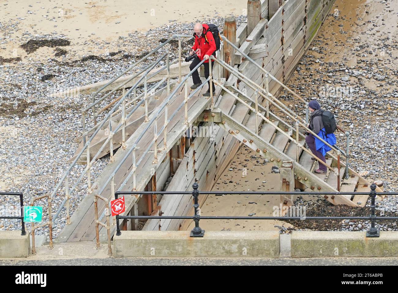 Vue aérienne de la plage de bord de mer couple marchant monter un petit escalier au-dessus de la sortie de l'aine du brise-lames jusqu'à la promenade verte rouge promeneur de chiens panneaux Cromer Norfolk UK Banque D'Images