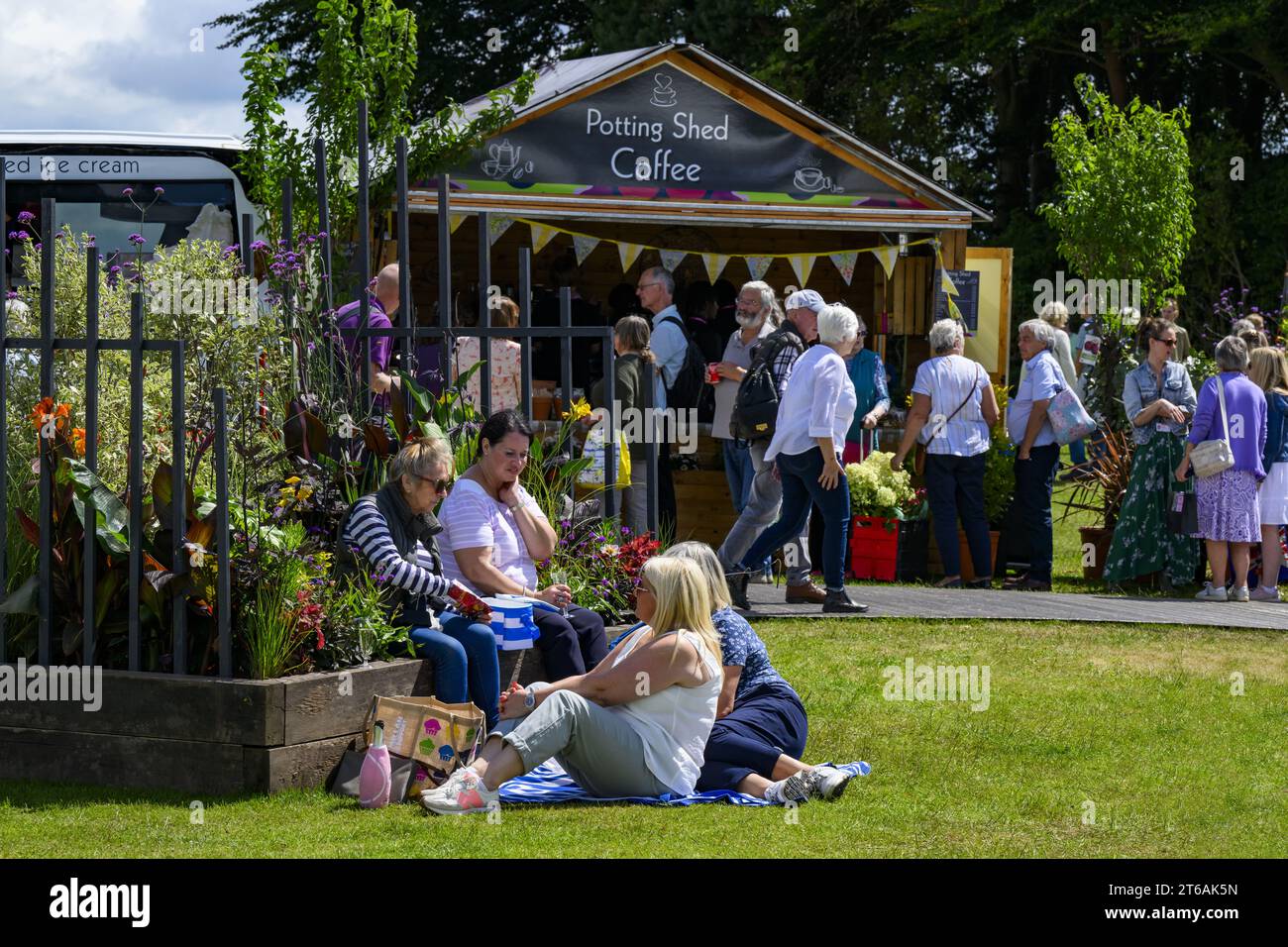 Visiteurs passant devant un stand de café et 4 quatre pique-niqueurs assis sur la pelouse en herbe au showground ensoleillé animé - 2023 Flower Show Tatton Park, Cheshire Angleterre Royaume-Uni. Banque D'Images