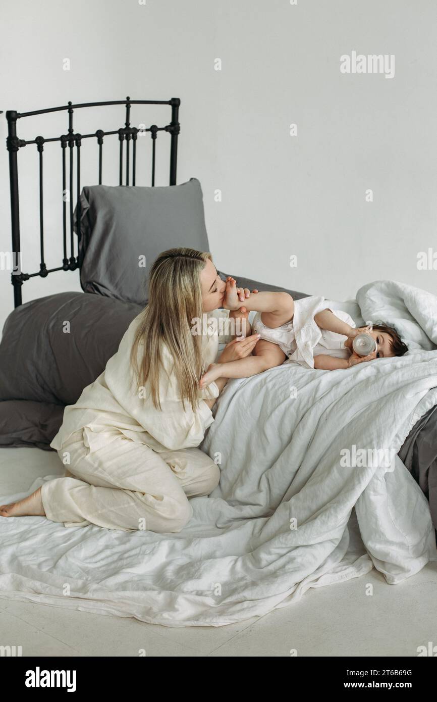 Une petite fille s'allonge sur le lit et boit de l'eau dans une bouteille, à côté de sa mère lui embrasse les pieds. Concept de famille. Vue à travers les barres métalliques du b Banque D'Images