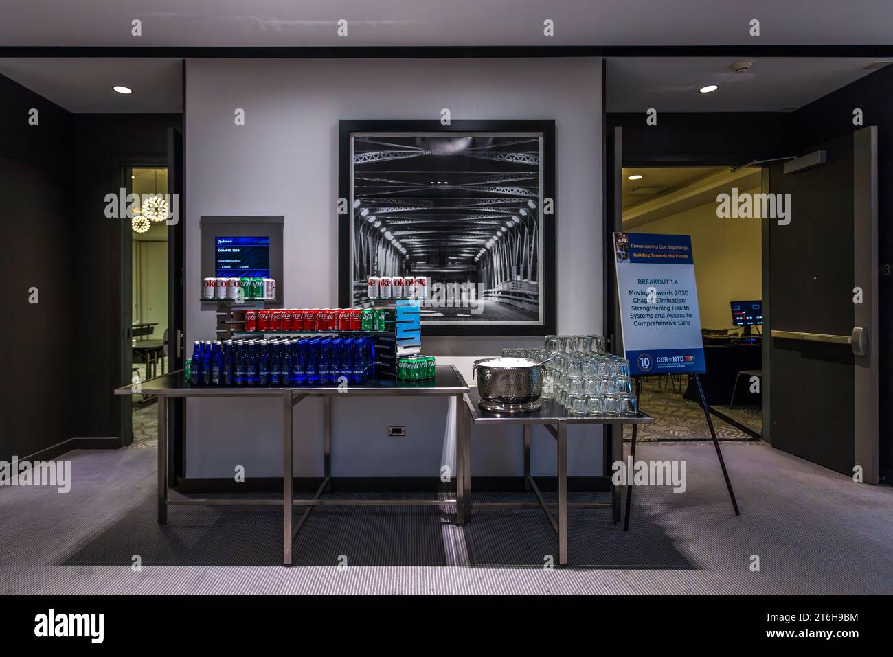 Une table de boissons avec des canettes et des bouteilles se trouve dans la zone de conférence du Radisson Blu Aqua Hotel à Chicago, Illinois, États-Unis, une grande photographie en noir et blanc d'un pont à treillis en acier est suspendue au-dessus, une pancarte à côté annonçant la session "Breakout 1.4 : Vers 2030 – Élimination de Chagas, Renforcement des Systèmes de Santé et Accès à des Soins Complets" d'une conférence internationale sur les maladies tropicales négligées. L'hôtel occupe les étages inférieurs de la tour Aqua, conçue en 2009 par l'architecte Jeanne Gang. Banque D'Images