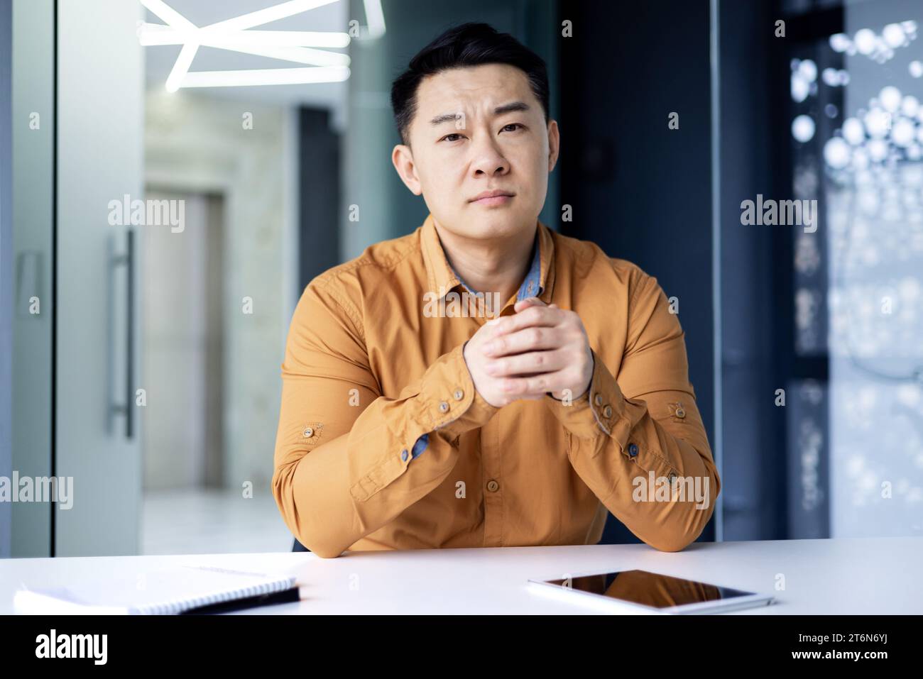 Portrait d'un travailleur de bureau asiatique sérieux assis à un bureau dans un bureau lumineux et parlant lors d'un appel vidéo, un homme chinois regardant la caméra avec les mains pliées, vue de la caméra. Banque D'Images
