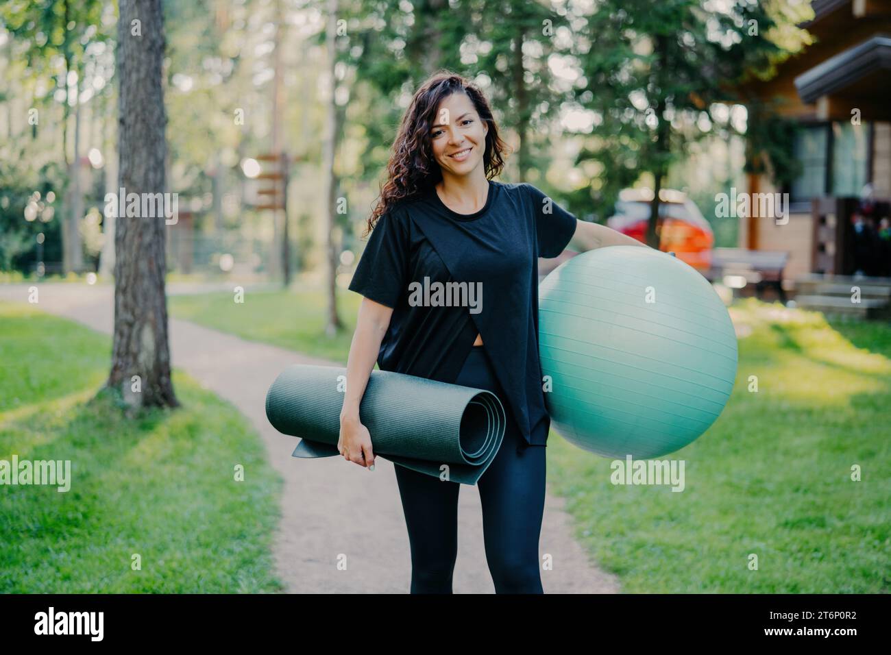 Femme souriante avec tapis de yoga et ballon d'exercice prêt pour une séance d'entraînement dans un cadre de parc serein Banque D'Images