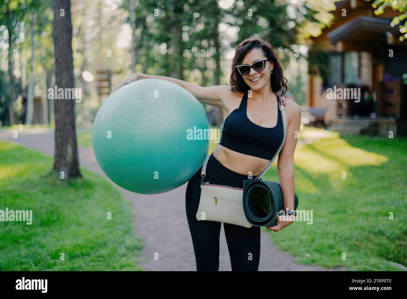 Femme active radieuse avec ballon d'exercice et tapis de yoga, arborant un sourire ensoleillé dans un parc luxuriant Banque D'Images
