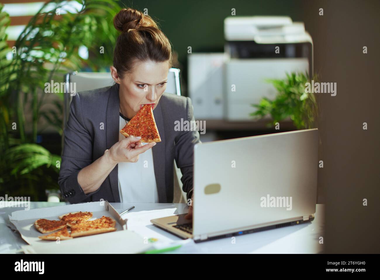Lieu de travail durable. femme moderne d'âge moyen travailleur dans un costume d'affaires gris dans le bureau vert moderne avec pizza et ordinateur portable. Banque D'Images