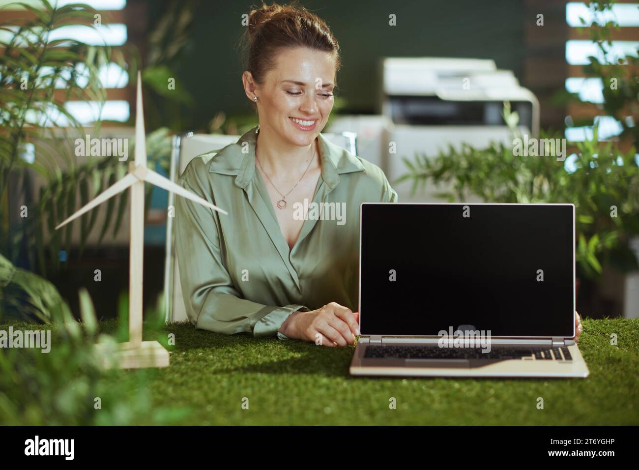 heureuse femme de propriétaire de petite entreprise moderne d'âge moyen en chemisier vert dans le bureau vert moderne avec moulin à vent montrant l'écran blanc d'ordinateur portable Banque D'Images