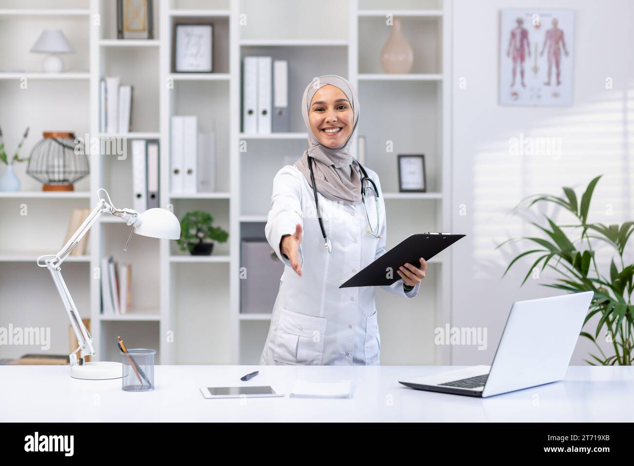 Portrait de femme médecin musulmane heureuse dans le hijab debout à table dans le bureau de l'hôpital, portant un manteau blanc, dossier dans les mains, étendant la main dans l'accueil, souriant et regardant la caméra. Banque D'Images