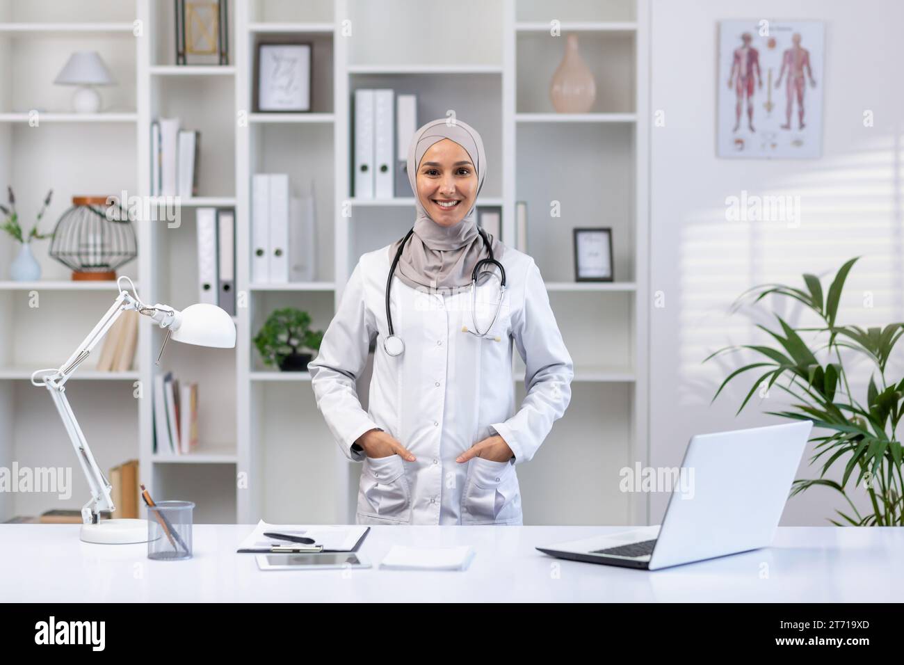 Portrait de femme médecin arabe heureuse dans le hijab debout à table dans le bureau de l'hôpital avec stéthoscope autour du cou, les mains dans les poches, souriant et regardant la caméra. Banque D'Images