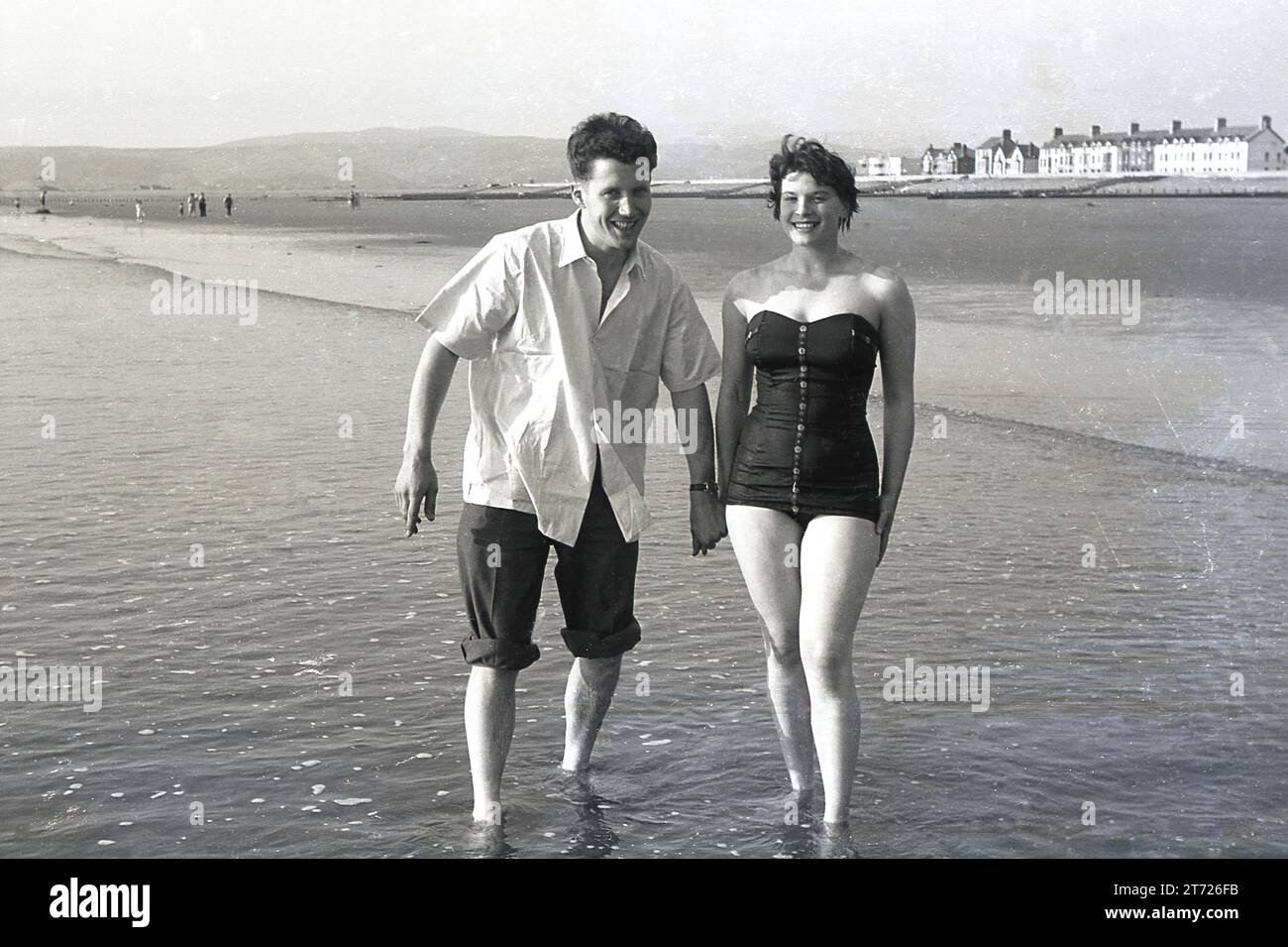 Années 1960, historique, un jeune couple adulte debout dans l'eau de mer à côté d'une plage se tenant par la main, Angleterre, Royaume-Uni. La jeune femme est en maillot de bain, tandis que le jeune mn a son pantalon enroulé, une chose masculine traditionnelle à faire au bord de la mer. Banque D'Images