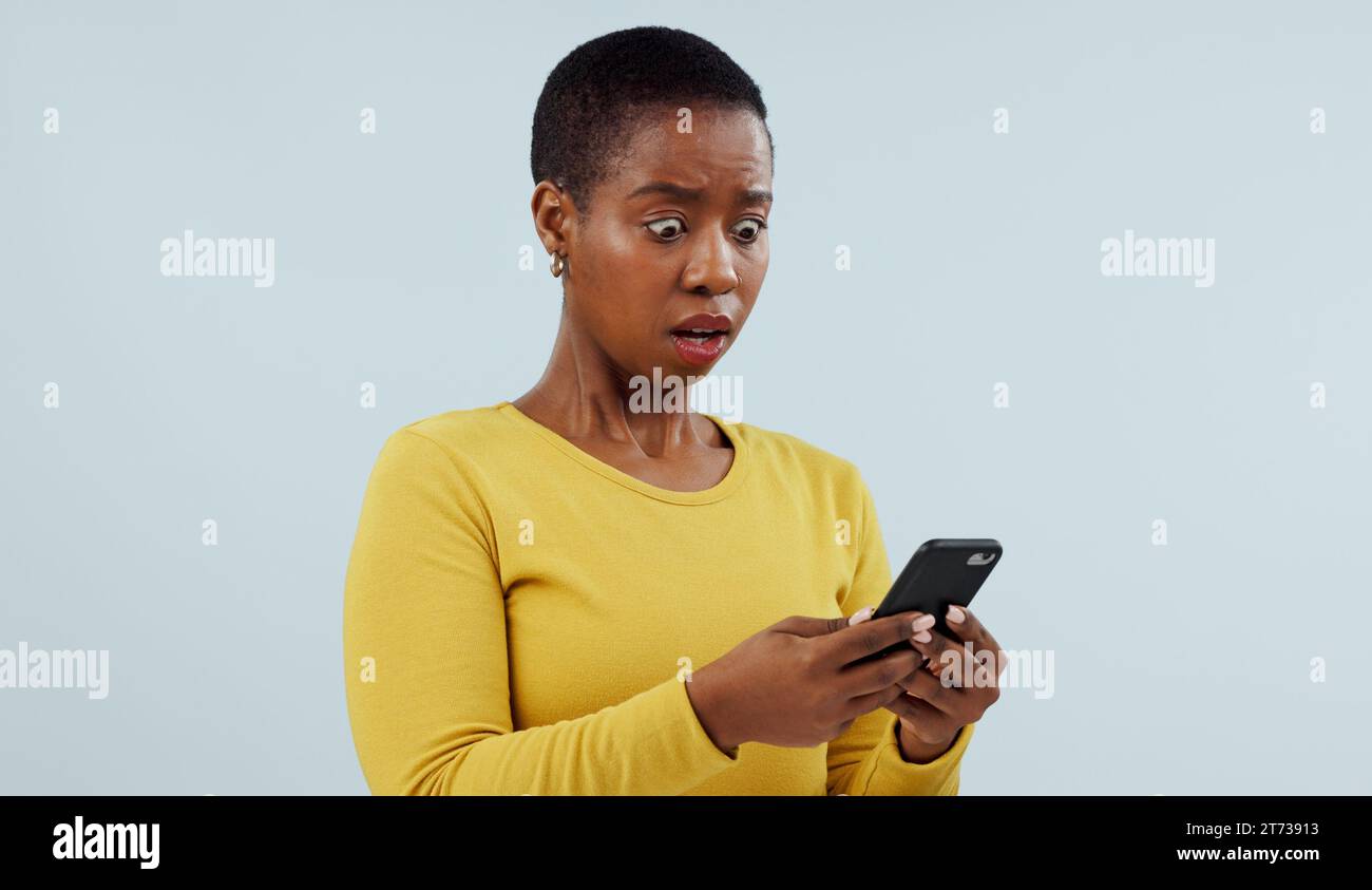 Femme noire, téléphone et nouvelles choquantes sur les réseaux sociaux, terrifiantes ou en ligne contre un fond de studio. Visage de la femme africaine surprise ou Banque D'Images