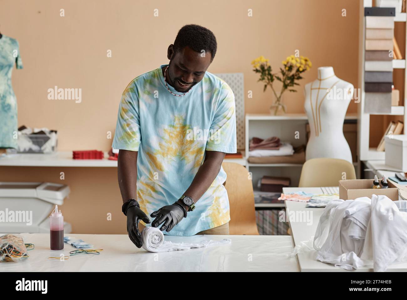 Jeune homme souriant dans des gants de protection roulant t-shirt blanc sur la table avant d'utiliser un colorant spécial pour créer un tout nouveau vêtement en studio Banque D'Images