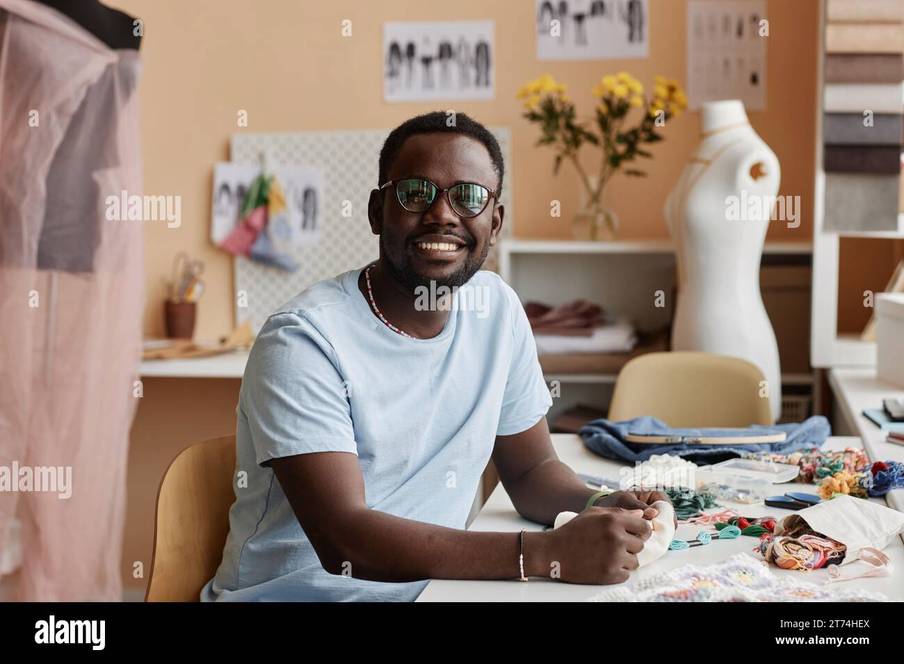 Jeune designer masculin afro-américain souriant dans des lunettes assis par la table avec des fournitures de couture dans l'atelier et regardant la caméra Banque D'Images