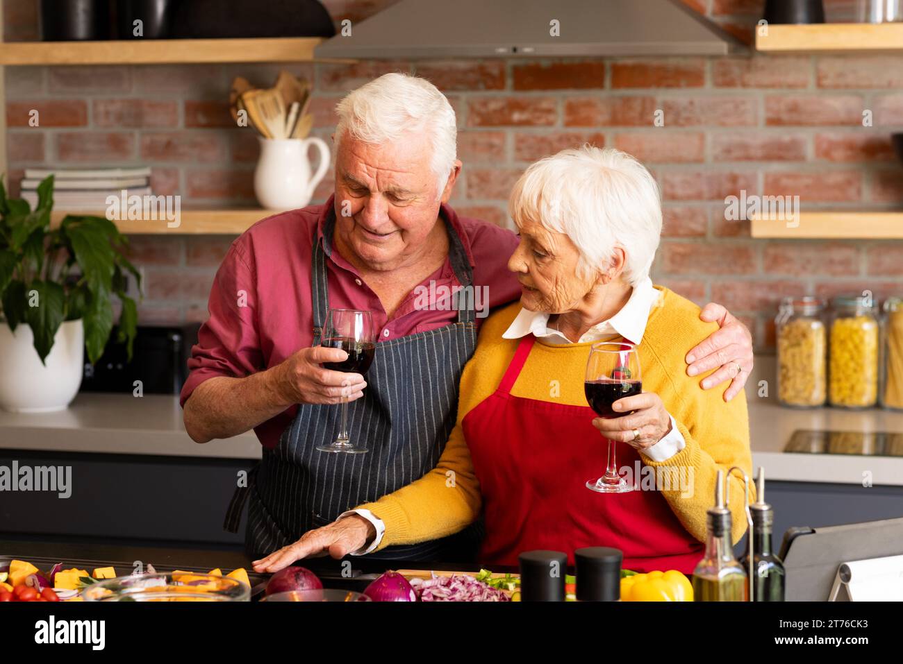 Heureux couple aîné caucasien préparant des légumes, buvant du vin et embrassant dans la cuisine Banque D'Images