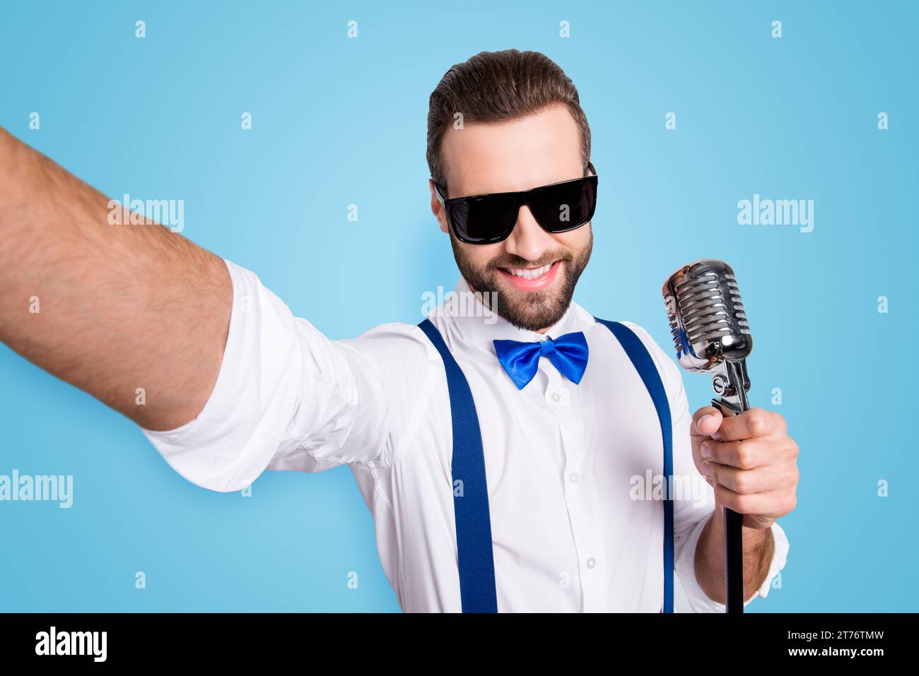 Autoportrait d'un chanteur élégant et élégant portant des bretelles de chemise en noeud papillon filant selfie sur l'appareil photo avant tenant le micro isolé sur fond gris Banque D'Images