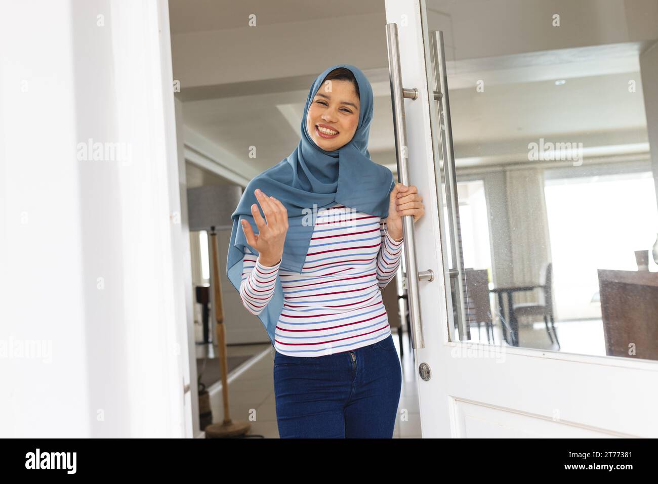 Heureuse femme biracial en hijab bleu debout à la porte d'entrée de la maison Banque D'Images