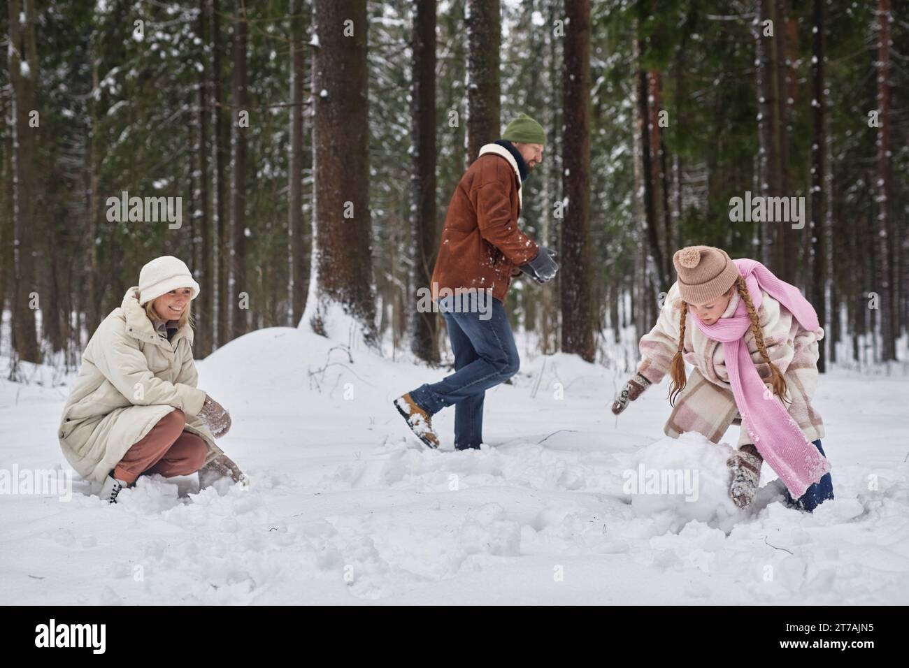 Jeune mère et fille roulant des boules de neige dans la forêt d'hiver ou le parc tandis que l'homme en vêtements d'hiver se déplaçant dans la dérive de neige derrière eux Banque D'Images
