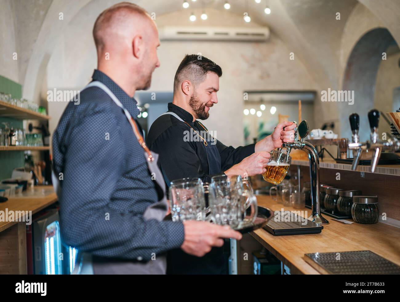 Deux hommes caucasiens amis barman bière tapant et serveur avec plateau habillé uniforme noir au comptoir de bar. Les gens au travail, le travail d'équipe, l'amitié des hommes et Banque D'Images