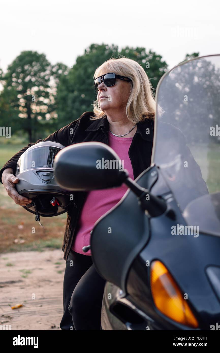 Portrait vertical de blonde mature en veste avec casque dans ses mains debout près de moto en automne dans la nature. Le tourisme moto. Une vie saine Banque D'Images