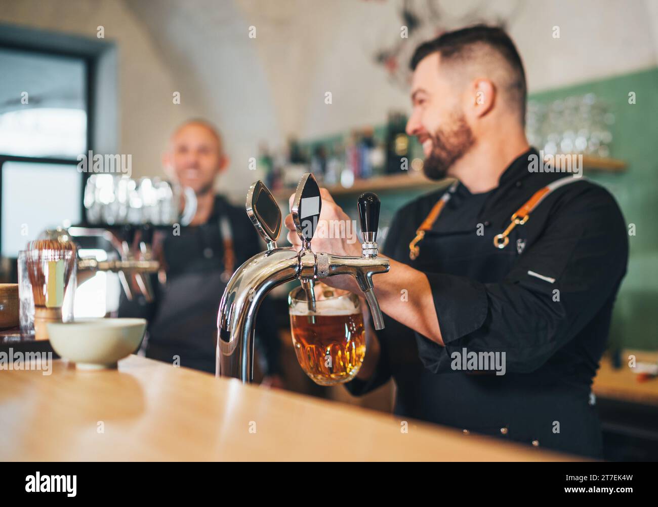 Tasse en verre de bière fraîche de lager et uniforme de barman vêtu souriant au serveur de coéquipier avec la bière de plateau tapant au comptoir de bar. Travail d'équipe réussi, fr Banque D'Images