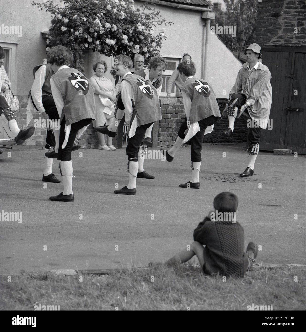 Années 1970, historlcal, un petit garçon assis regardant des hommes adultes et des jeunes hommes vêtus de costumes faisant une danse morris, Angleterre, Royaume-Uni. La danse Morris est une forme populaire de danse folklorique anglaise jouée lors de festivals et de vacances pour célébrer l'arrivée de l'été et la récolte d'or de l'automne. Banque D'Images