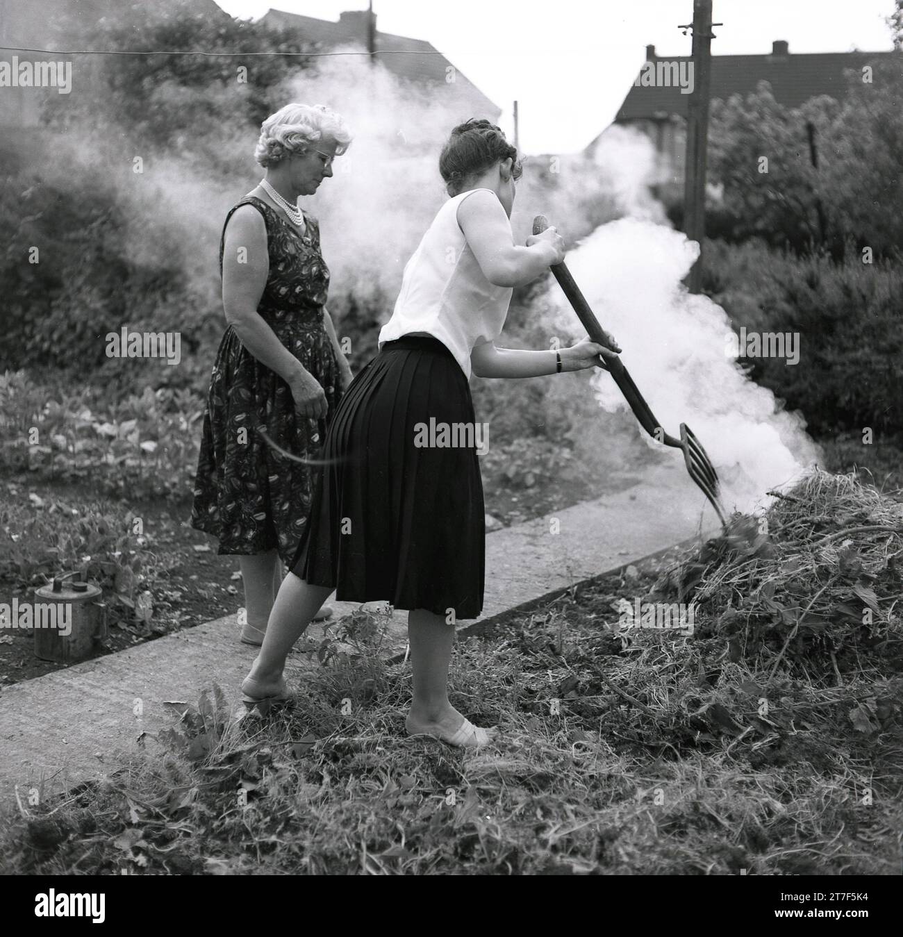 Années 1960, historique, deux dames brûlant de la végétation sur un feu de joie dans un jardin arrière, une dame dans un haut sans manches et une jupe plissée, utilisant une fourchette de jardin pour ajouter des feuilles au feu, Angleterre, Banque D'Images