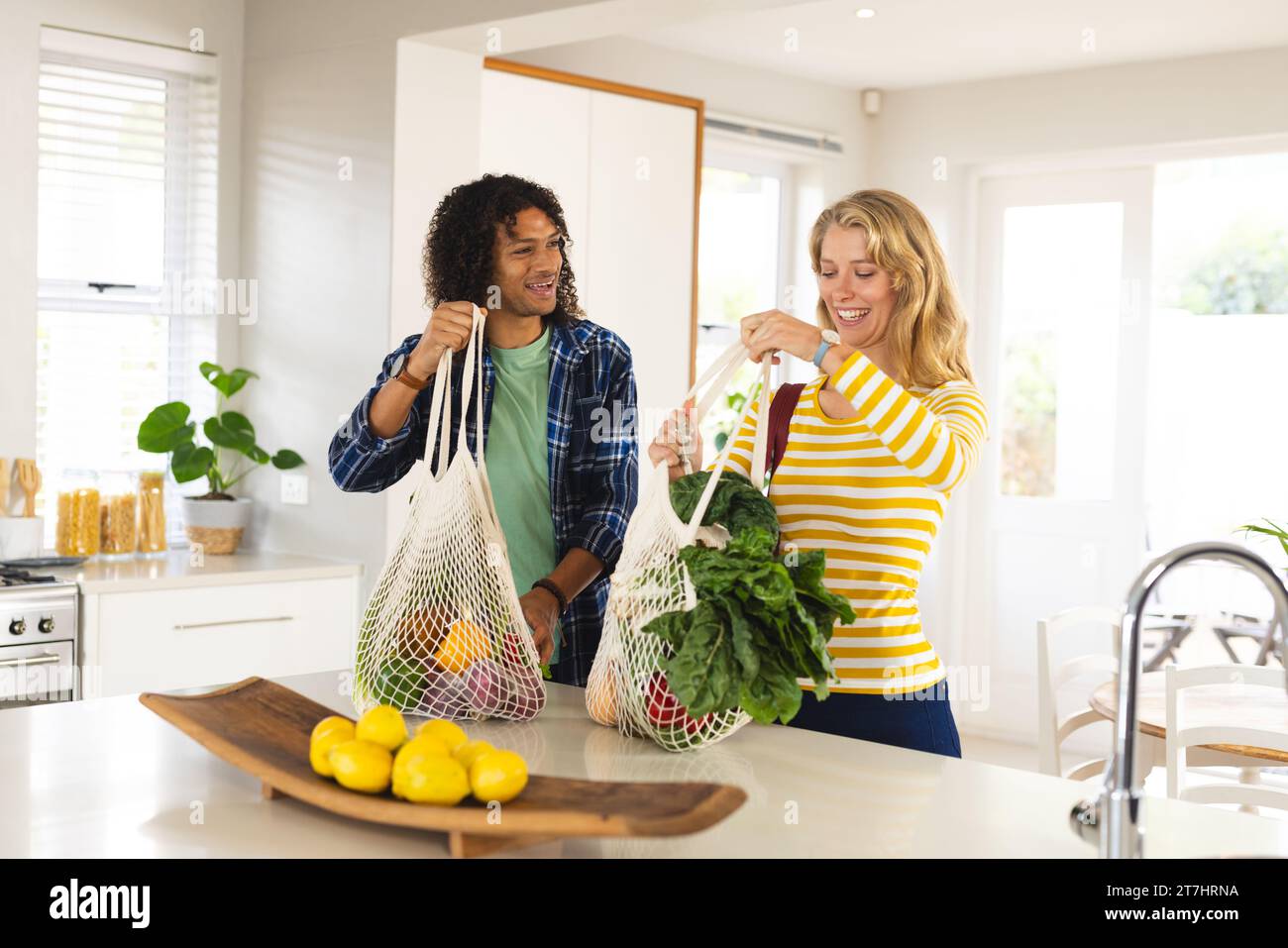 Heureux couple diversifié déballant l'épicerie et souriant dans la cuisine ensoleillée Banque D'Images