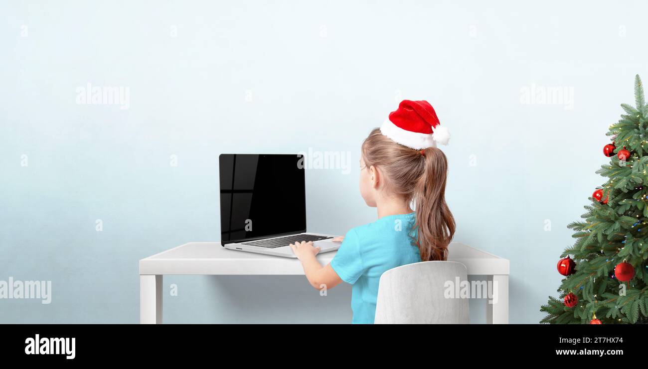 Fille avec chapeau de Noël rouge travaille sur ordinateur portable dans la chambre avec arbre de vacances. Soncept d'écrire des cartes de voeux et des lettres au Père Noël Banque D'Images