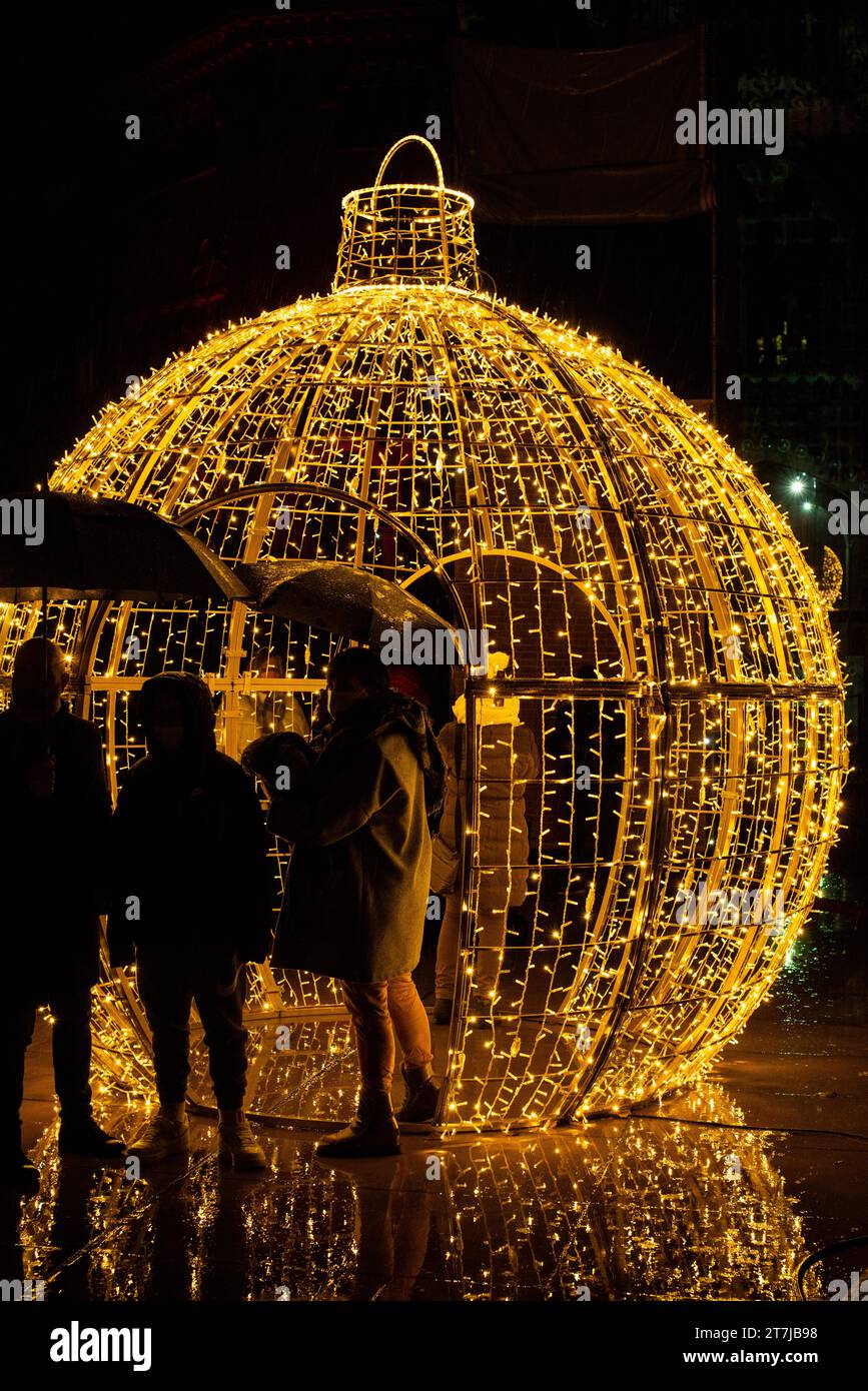 Sous la lueur festive, les silhouettes se mêlent à la magie de la nuit à côté d'une boule de Noël géante, créant une scène charmante remplie de vacances Banque D'Images