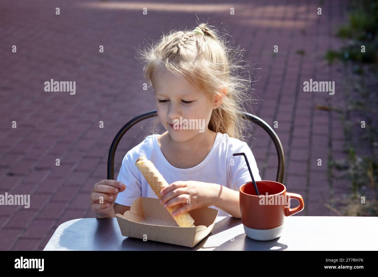 Petite fille souriante prendre un petit déjeuner dans un café d'été en plein air. Mignonne fille caucasienne mangeant un tube à gaufres savoureux et buvant du cacao Banque D'Images