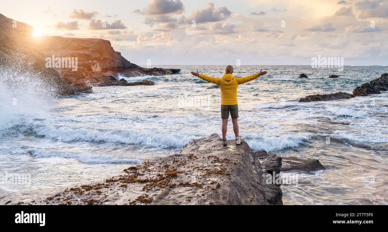 Personne en veste jaune debout sur le rocher au bord de la mer avec les bras grands ouverts au coucher du soleil Banque D'Images