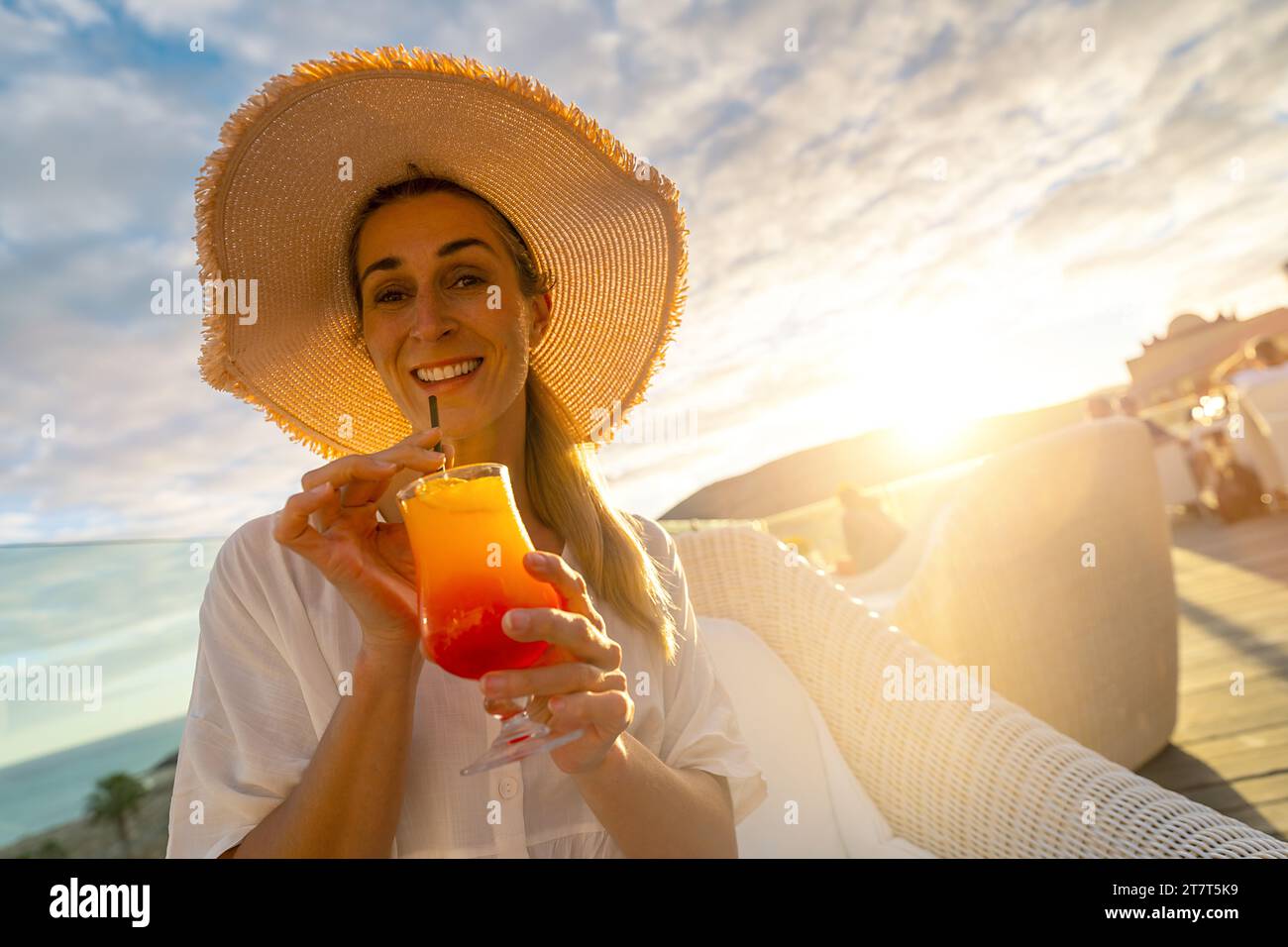Femme en chapeau de paille sirotant un cocktail tropical à l'hôtel tropical de plage sur le coucher du soleil avec un fond flou Banque D'Images