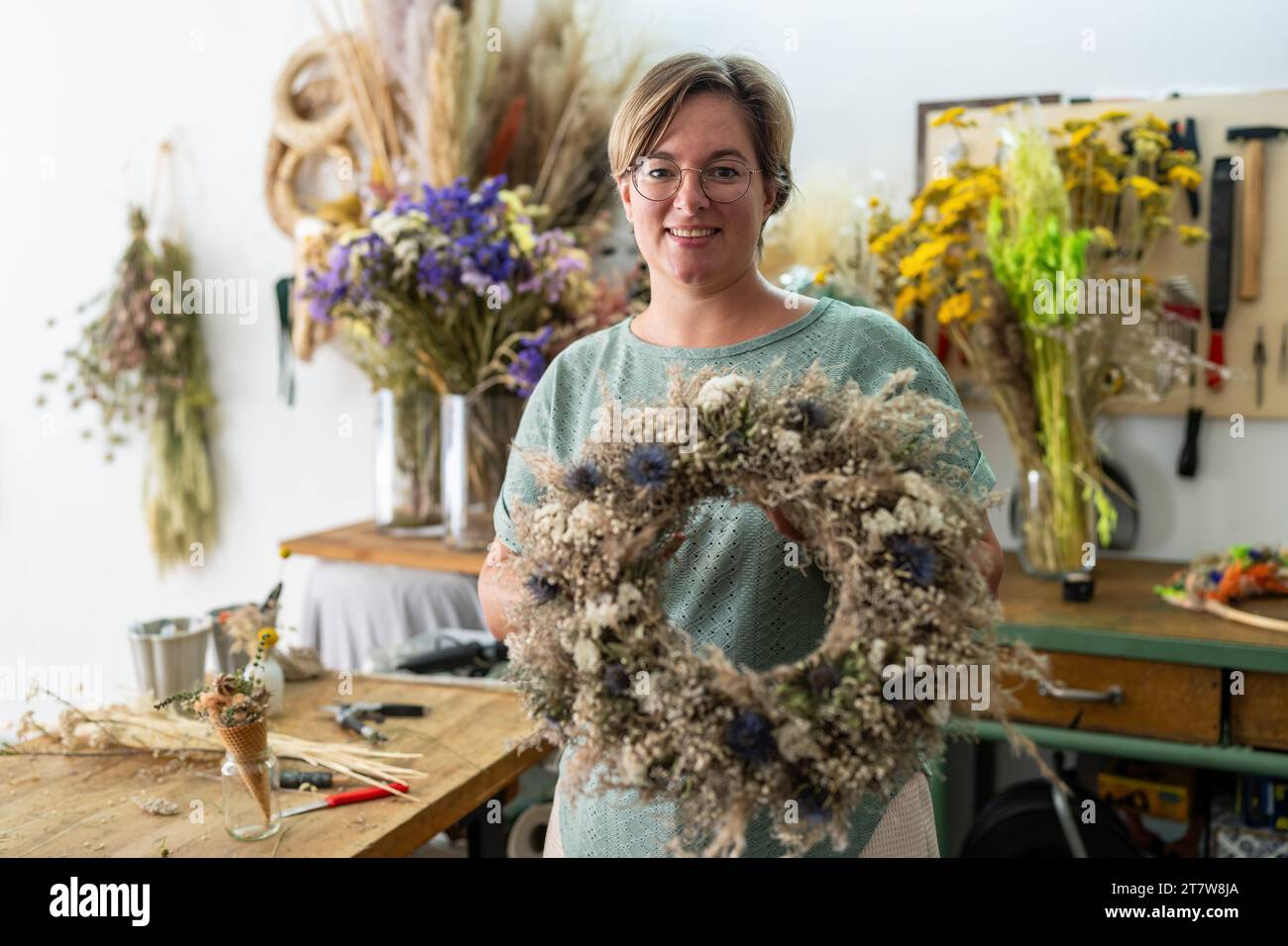 Femme avec des lunettes souriant et tenant une grande couronne florale séchée dans un atelier d'artisanat Banque D'Images