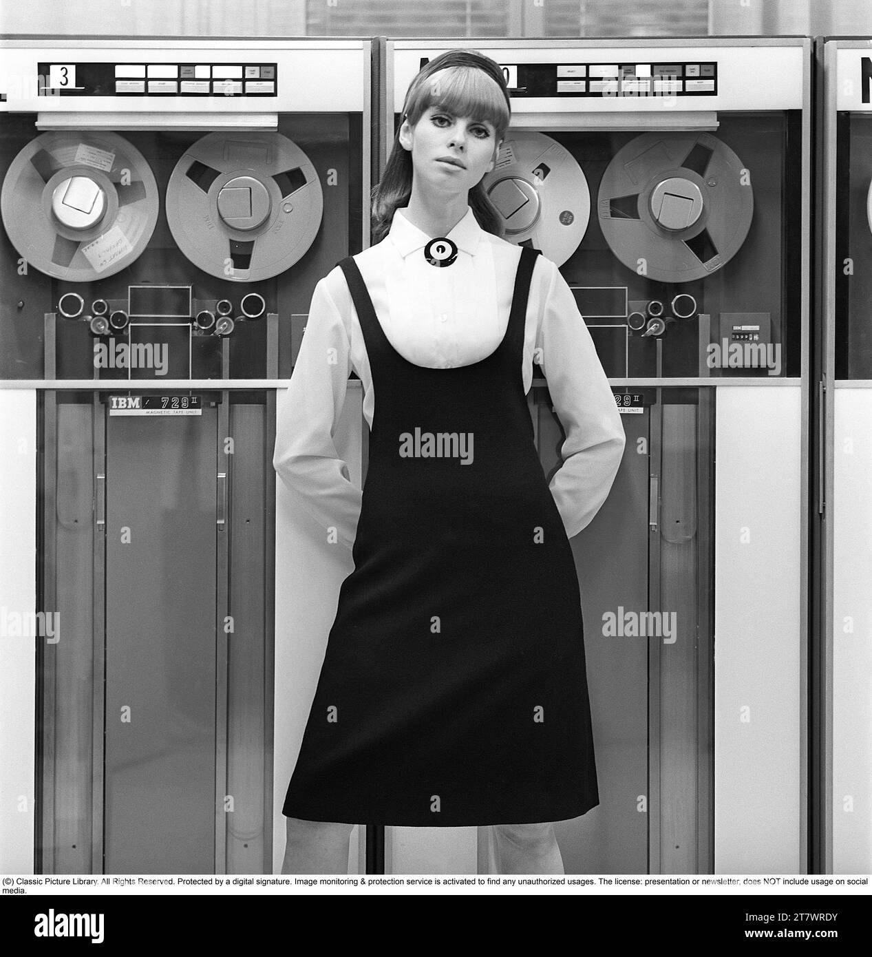 Dans les années 1960 Mannequin portant les vêtements typiques de 1965. La photographie est prise dans une salle informatique centrale des années 1960 avec des unités de bande magnétique utilisées où la bande contenant des informations est lue ou entrée. Elle porte une chemise blanche et une robe noire par-dessus. Photo prise 1965. Réf. DV16 Banque D'Images