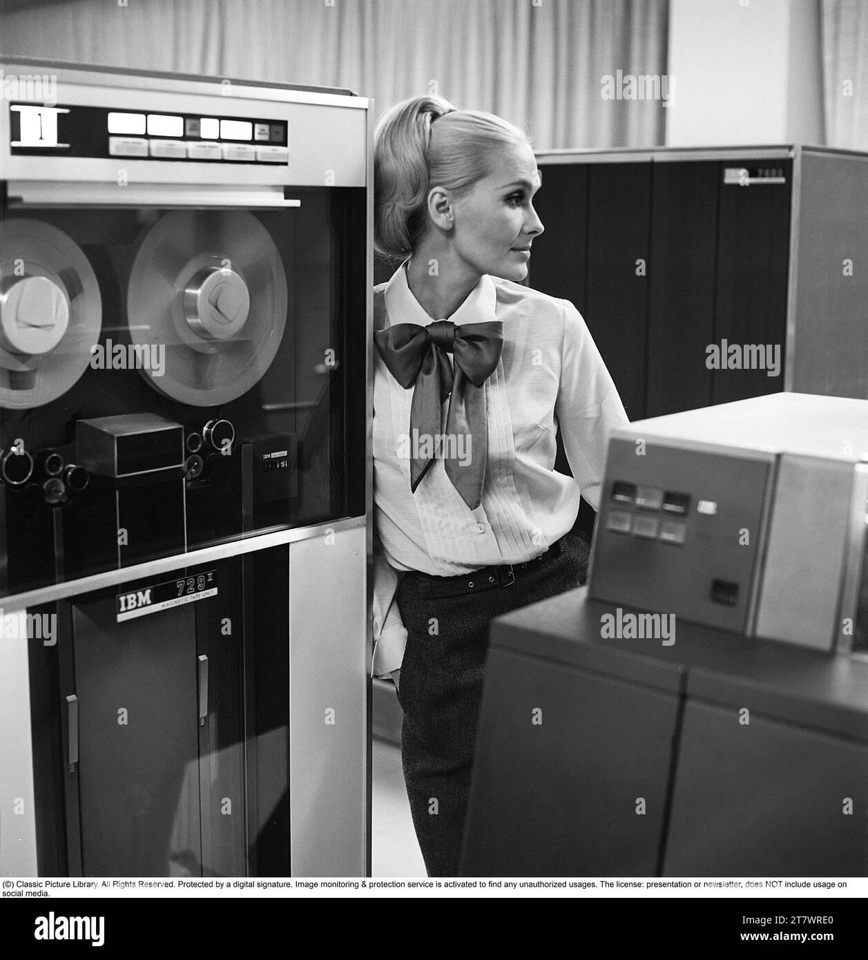 Dans les années 1960 Mannequin portant les vêtements typiques de 1965. La photographie est prise dans une salle informatique centrale des années 1960 avec des unités de bande magnétique utilisées où la bande contenant des informations est lue ou entrée. Elle porte une chemise blanche. Photo prise 1965. Réf. DV16 Banque D'Images