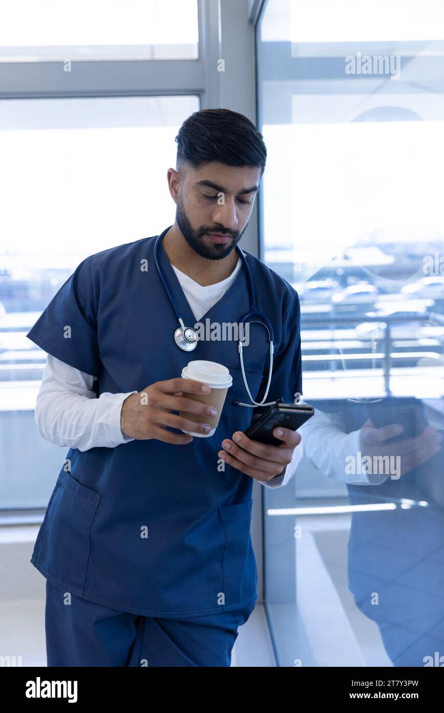 Médecin masculin biracial utilisant smartphone et tenant une tasse de café dans le bureau de l'hôpital Banque D'Images