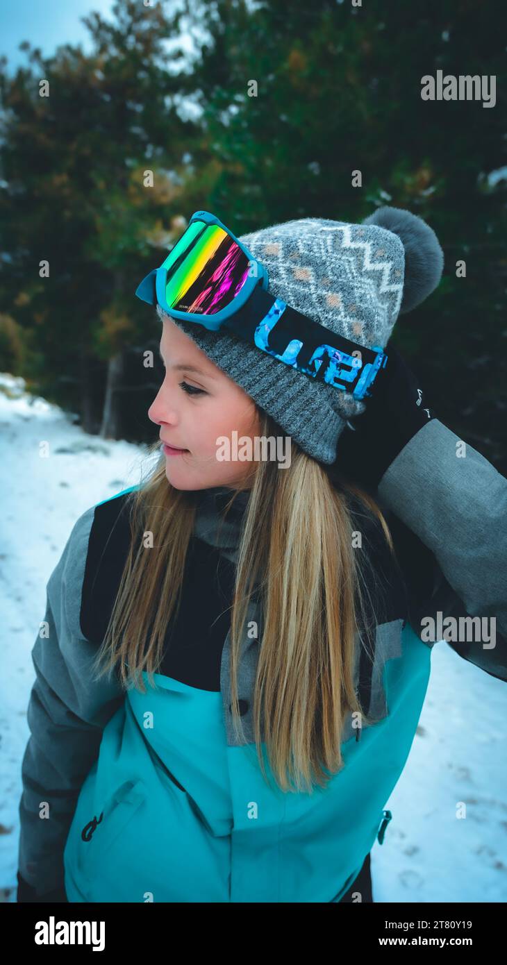 Une jeune skieuse souriante se tient dans la neige, vêtue d'un manteau d'hiver et de lunettes, tenant un snowboard sur son épaule Banque D'Images