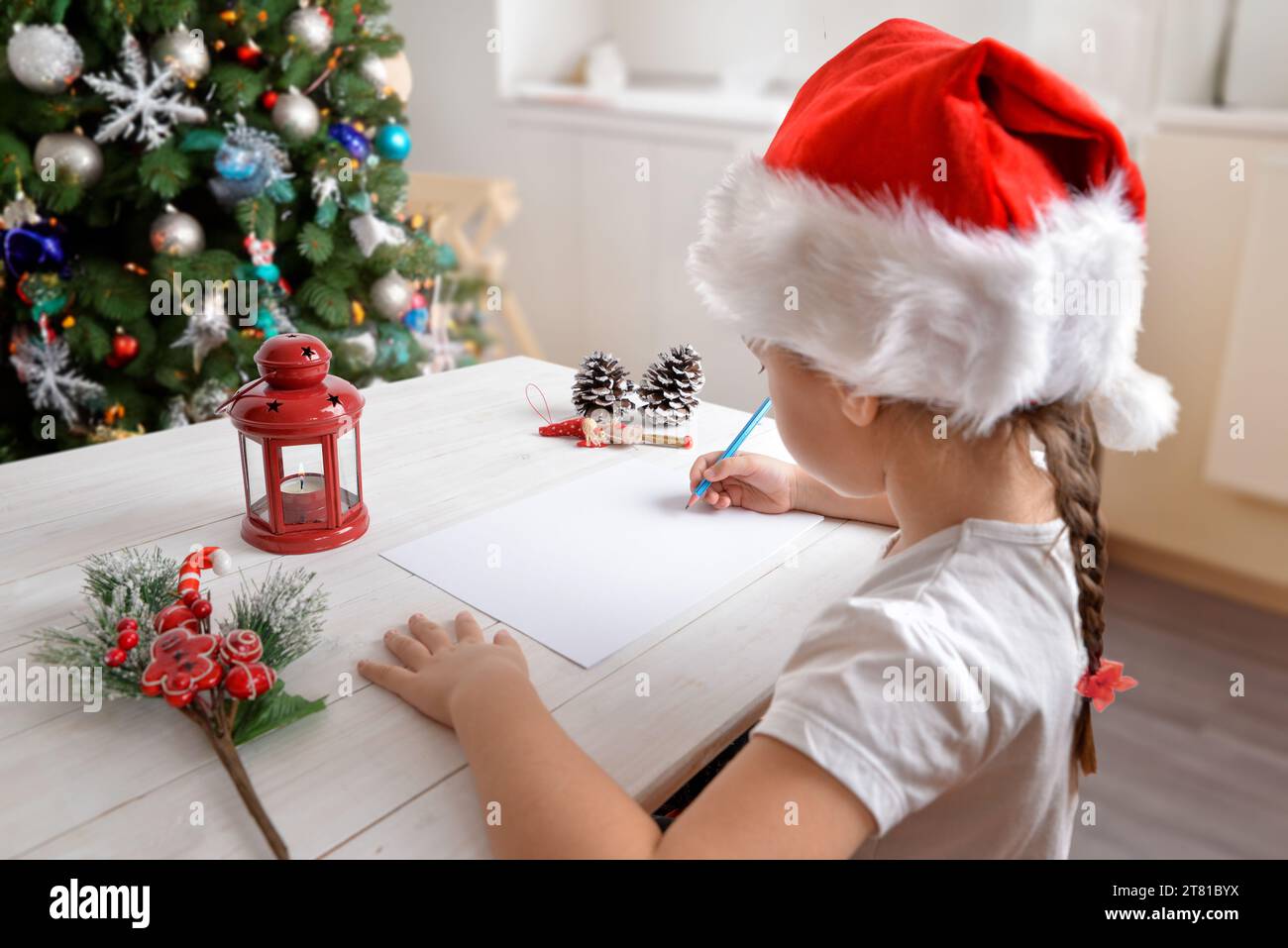 Fille écrit lettre au Père Noël à la maison avec chapeau de Noël. Sapin et décorations festives en arrière-plan. Scène de vacances réconfortante Banque D'Images